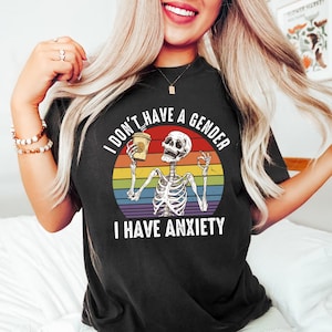Puede incluir: Camiseta negra con un gráfico de estilo vintage de un esqueleto sosteniendo una taza de café. El gráfico incluye el texto "No tengo género Tengo ansiedad" y una franja de arcoíris.