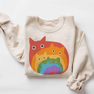 May include: A beige sweatshirt featuring a rainbow design of cats of varying sizes. The largest cat is red, followed by orange, then yellow, green, blue, and purple.