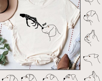 May include: A white t-shirt with a black skeleton hand reaching out to touch a dog's nose. The text "BOOP" is above the dog's head. The shirt is a graphic tee with a cute and funny design.