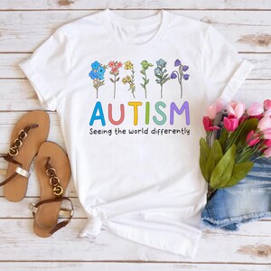 May include: White t-shirt with a rainbow of flowers and the word "AUTISM" in rainbow colors. The text below reads "Seeing the world differently".