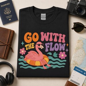 May include: A black t-shirt featuring a graphic of a pink flamingo wearing heart-shaped sunglasses while floating in an orange and yellow inner tube on teal waves, with the text "GO WITH THE FLOW" in orange, pink, and purple retro-style font.
