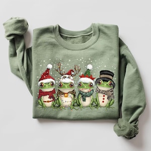 May include: A green sweatshirt with a festive design featuring four frogs wearing Christmas hats and accessories. The frogs are lined up in a row and are surrounded by snowflakes.
