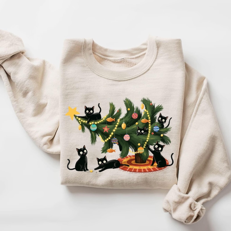 Op de afbeelding: Een beige sweatshirt met een kerstontwerp. Het heeft een kerstboom versierd met ballen en lichtjes, omringd door zwarte katten. De katten spelen met de boom, wat een speelse, feestelijke sc&egrave;ne cre&euml;ert.
