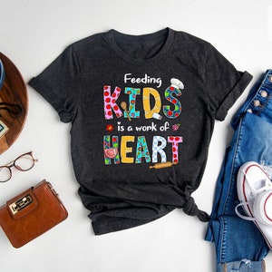 May include: A dark gray t-shirt with the text "Feeding Kids is a work of Heart" in colorful patchwork letters. The letters are decorated with images of kitchen utensils and food.