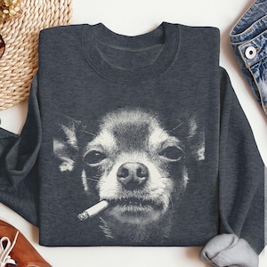 Op de afbeelding: Donkergrijze sweatshirt met een zwart-wit afbeelding van een Chihuahua met een sigaret in zijn mond. Het gezicht van de hond is het middelpunt, met gedetailleerde vacht en een enigszins samengeknepen uitdrukking. De sweatshirt heeft een ronde hals en lange mouwen.