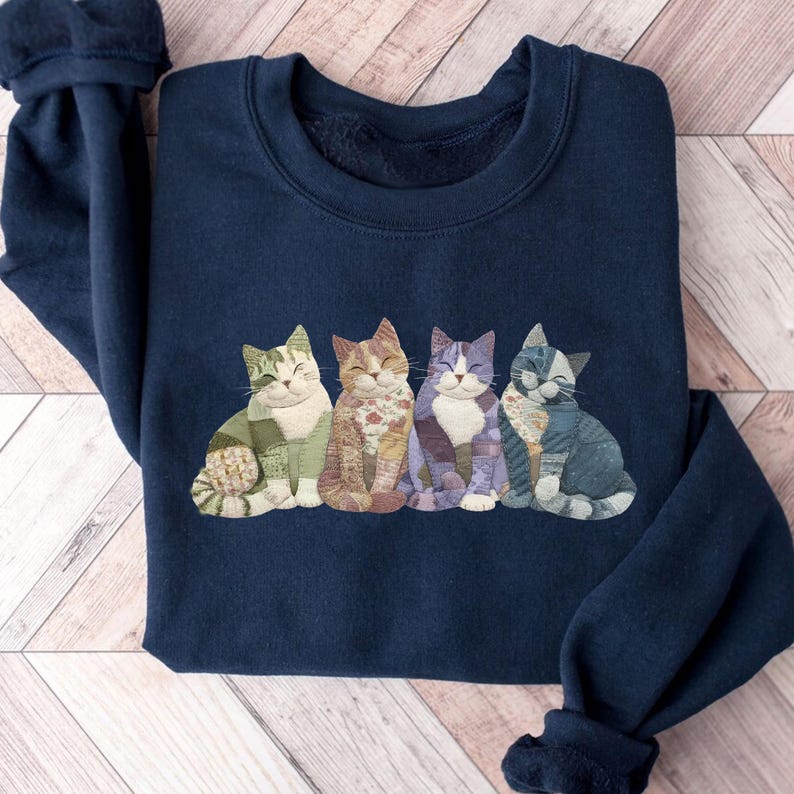 Peut inclure: Sweat-shirt bleu marine avec quatre illustrations de chats stylis&eacute;s. Chaque chat pr&eacute;sente un motif patchwork unique dans les tons de vert, marron, violet et bleu. Col rond et manches longues.