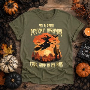 Może przedstawiać: Oliwkowozielony t-shirt z grafiką przedstawiającą czarownicę lecącą na miotle nad pustynnym krajobrazem o zachodzie słońca. Napis ON A DARK DESERT HIGHWAY oraz COOL WIND IN MY HAIR wykonano w postarzanej żółtej czcionce, z widocznymi kaktusami i nietoperzami.