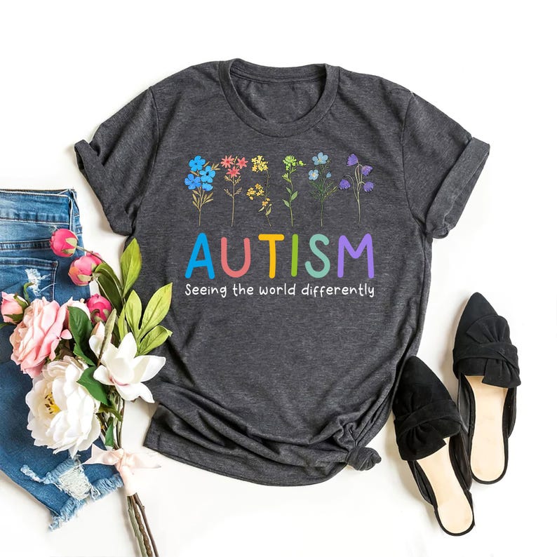 May include: A dark gray t-shirt with a rainbow-colored "AUTISM" graphic and the text "Seeing the world differently". The graphic features a row of colorful flowers.