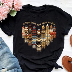 May include: Black t-shirt with a graphic of colorful feathers and the text "Feathers appear when angels are near".