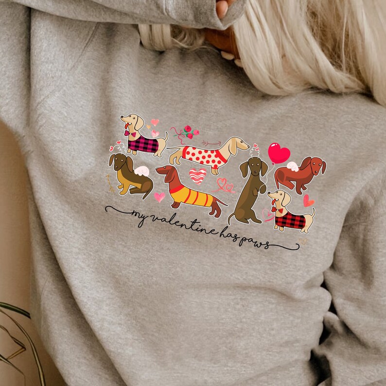 Puede incluir: Una sudadera gris jaspeado con un estampado gr&aacute;fico de varios perros salchicha con jers&eacute;is y pajaritas, rodeados de corazones rojos y rosas y el texto "my valentine has paws" escrito en una fuente cursiva de color negro.