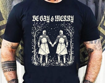May include: Black t-shirt with a white graphic of two knights holding hands in a forest setting. The text "BE GAY & MERRY" is printed above the graphic.