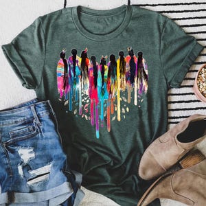 May include: A green t-shirt with a colorful heart-shaped design featuring a group of women with long hair and feathers. The design is reminiscent of Native American culture.