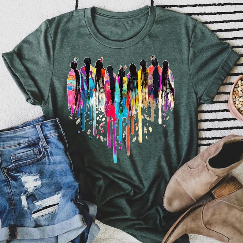 May include: A green t-shirt with a colorful heart-shaped design featuring a group of women with long hair and feathers. The design is reminiscent of Native American culture.