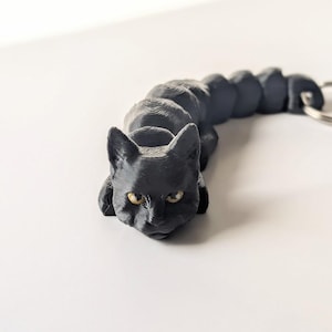May include: A black 3D printed cat keychain with yellow eyes. The cat is curled up in a sleeping position.