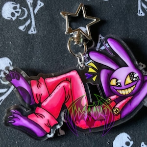 May include: Purple and pink cartoon character with a large grin and yellow eyes. The character is lying on its back with its legs in the air. The character is attached to a silver star-shaped keychain.