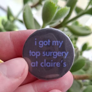 May include: A circular, dark grey badge with the purple text "i got my top surgery at claire's". The badge is held in a hand, with a blurred green plant in the background.
