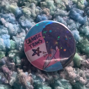 May include: A round button with a glittery, holographic finish. The button features a cartoon illustration of a person with a transgender pride flag behind them. The text "CHAOTIC TRANS" is written on the flag.