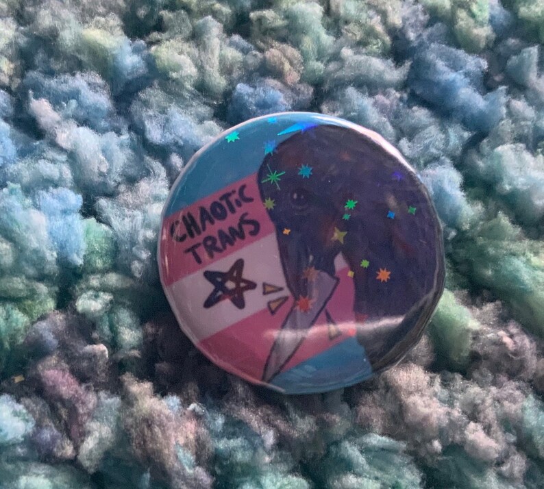 May include: A round button with a glittery, holographic finish. The button features a cartoon illustration of a person with a transgender pride flag behind them. The text "CHAOTIC TRANS" is written on the flag.
