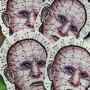 May include: Four stickers of a man's face with a grid of red lines and black pins over his face. The man has a serious expression and is looking straight ahead.