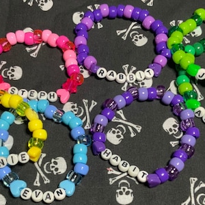 May include: A collection of colorful beaded bracelets with names spelled out in white letter beads. The bracelets are in shades of pink, purple, green, yellow, and blue. The background is a black fabric with a white skull and crossbones pattern.