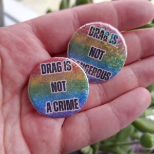 May include: Two round buttons with rainbow backgrounds and the text "DRAG IS NOT A CRIME" and "DRAG IS NOT DANGEROUS". The buttons are held in a hand, with green foliage in the background.