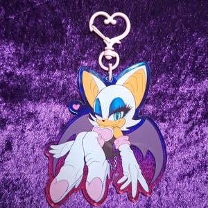 May include: A pink and blue acrylic keychain featuring a cartoon bat character with white, blue, and orange details. The keychain has a heart-shaped clasp and a glittery pink border. The character is in a relaxed pose.