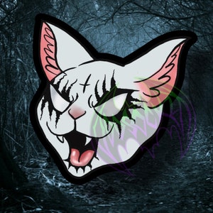 Op de afbeelding: Een witte kat met zwarte markeringen rond de ogen en de mond, met een kruis op het voorhoofd. De kat heeft een dreigende uitdrukking en wordt getoond tegen een donkere, schaduwrijke achtergrond.