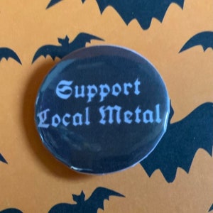 Może przedstawiać: Okrągły guzik w kolorze czarnym i białym z napisem "Support Local Metal" w białym gotyckim kroju pisma.