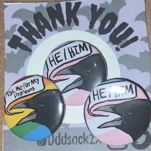 May include: Three round buttons with a black crow design. The buttons have the text "Ask Me For My Pronouns", "He/Him", and "He/Him" on them. The buttons are on a purple background with the text "Thank You!" on the top.
