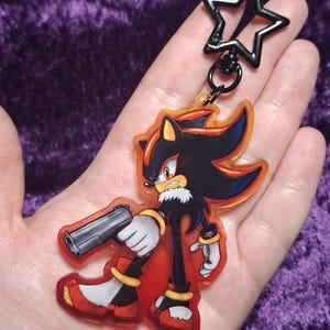 May include: A clear acrylic keychain featuring the character Shadow the Hedgehog from the Sonic the Hedgehog franchise. Shadow is depicted in a dynamic pose, holding a gun. The keychain has a black star-shaped clasp and a red and orange border.