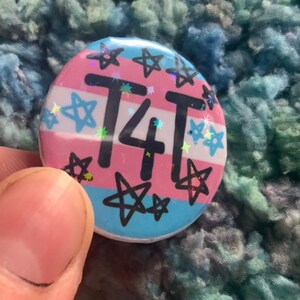 May include: A round button with a transgender pride flag design. The button has a pink, blue, and light blue background with white stars and the text "T4T" in black.