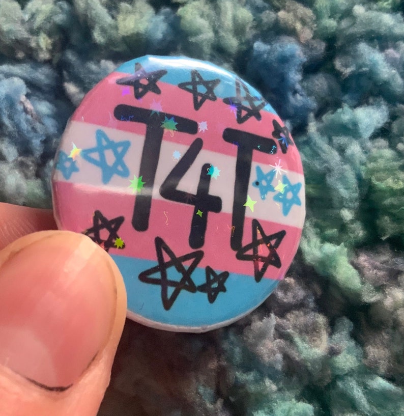May include: A round button with a transgender pride flag design. The button has a pink, blue, and light blue background with white stars and the text "T4T" in black.