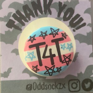 May include: A round button with a pink, blue, and white transgender pride flag design. The button has black stars and the text "T4T" in black. The button is on a purple background with a white border and the text "THANK YOU!" in black. The text "@Oddsockzx" is in black at the bottom of the image.
