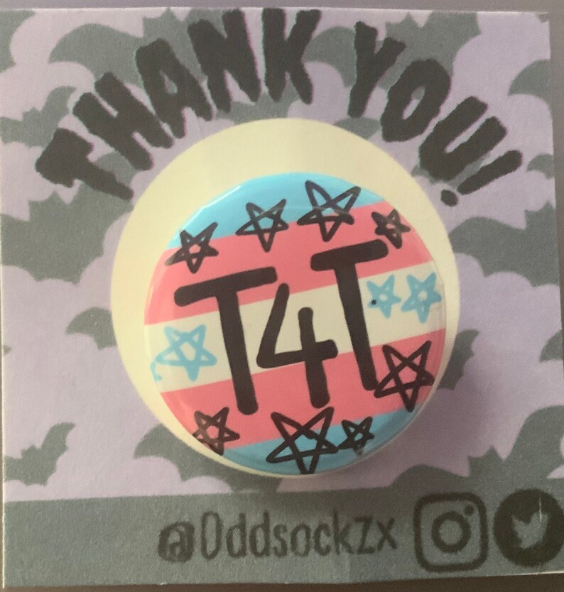 May include: A round button with a pink, blue, and white transgender pride flag design. The button has black stars and the text "T4T" in black. The button is on a purple background with a white border and the text "THANK YOU!" in black. The text "@Oddsockzx" is in black at the bottom of the image.
