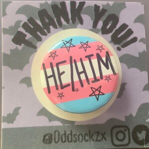May include: A pink, blue, and white button with the text "HE/HIM" in black. The button has a white border and is decorated with five-pointed stars.