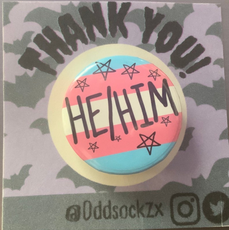 May include: A pink, blue, and white button with the text "HE/HIM" in black. The button has a white border and is decorated with five-pointed stars.