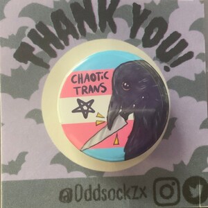 May include: A round button with a black crow holding a knife. The crow is depicted with a white eye and a pink beak. The button has a transgender pride flag design with the words "Chaotic Trans" and a pentagram. The button is on a purple background with bats.