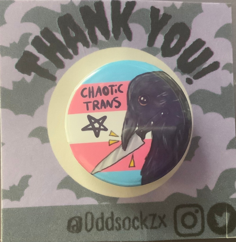 May include: A round button with a black crow holding a knife. The crow is depicted with a white eye and a pink beak. The button has a transgender pride flag design with the words "Chaotic Trans" and a pentagram. The button is on a purple background with bats.