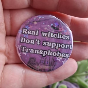Peut inclure: Un bouton rond violet avec le texte blanc "Real witches Don't support Transphobes". Le bouton présente un motif scintillant en forme d'étoiles et une silhouette de fioles de potion en bas.