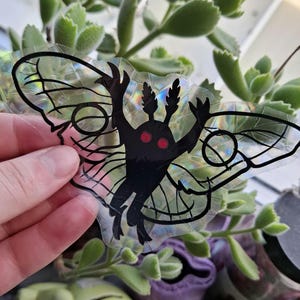 May include: A black and red moth sticker with iridescent wings. The moth has large, red eyes and is in a flying position.