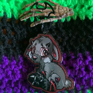 May include: A grey and red cartoon bunny with large eyes and a bloody nose. The bunny is wearing a red and black outfit and has glitter on its fur. The bunny is hanging from a skeleton hand.