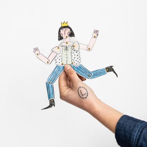 Puede incluir: Muñeco de papel dibujado a mano de un rey con corona dorada, pelo negro y atuendo a rayas blancas y azules. El muñeco tiene articulaciones y es sostenido por una mano sobre un fondo blanco.