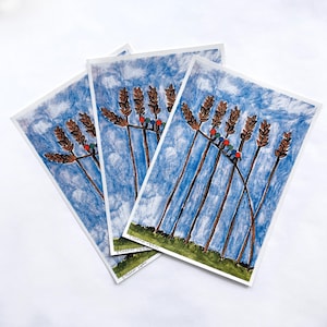 Puede incluir: Tres impresiones artísticas con una ilustración en acuarela de espigas de trigo sobre un cielo azul con nubes. El trigo es marrón y el cielo es azul y blanco. Pequeños puntos rojos y azules están en las espigas de trigo. Las impresiones son sobre papel blanco.