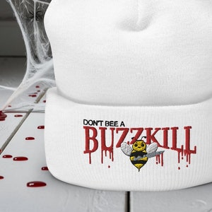 May include: White beanie hat featuring the embroidered text "DON'T BE A BUZZKILL" in red, with a cartoon bee holding a knife. The design is set against a Halloween backdrop with a spider web and fake blood.