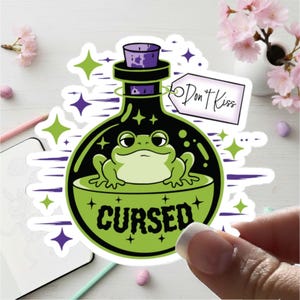 May include: A die-cut sticker featuring a green frog inside a round glass bottle with a purple stopper. The bottle has the word CURSED printed in black and a white tag attached that reads Don't Kiss. The design includes lime green and purple stars.