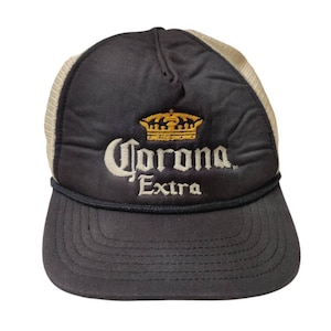 May include: A black and white trucker hat with a white mesh back. The front of the hat has a yellow embroidered crown and the text "Corona Extra".