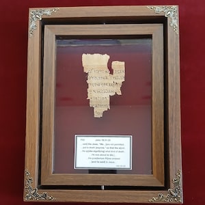 May include: Framed fragment of ancient text, likely a religious document, with visible script. The text is displayed within a double wooden frame with ornate corner accents, set against a red background. A small label with additional text is visible.