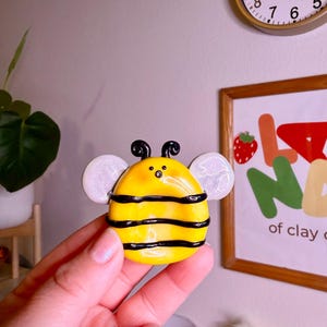 May include: A yellow and black bee-shaped ceramic figurine with white wings. The bee has black stripes and antennae. The figurine is held in a hand, with a clock and a framed picture in the background.