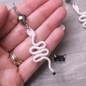 May include: White crocheted snake earrings with a gold and black crystal bead. The earrings are hanging from silver hooks.