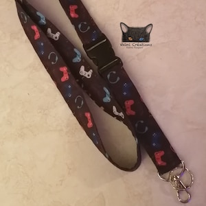 May include: A black lanyard patterned with colourful video game controllers and headphones. It has a black plastic buckle and a silver metal clasp. The image includes the text "Valni Creations".